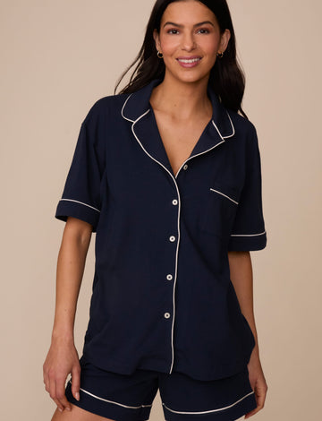 On a neutral studio backdrop, Marcela wears the Perfect Pima Pajama Shirt in Navy with matching navy pajama shorts. The front view highlights the relaxed fit, short sleeves, button-front closure, contrast piping, and chest pocket.  sizing: Marcela wears size Small