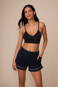 On a neutral studio backdrop, Marcela wears the Perfect Pima Pajama Short in Navy with a black bralette. The front view highlights the relaxed fit, soft elastic waistband, side pockets, and contrast piping along the hem. sizing: Marcela wears size Small