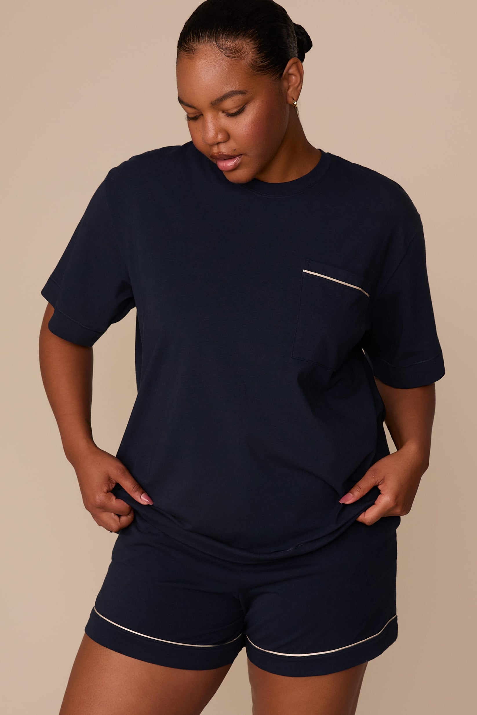 On a warm beige studio backdrop, Zarria wears the Perfect Pima Pajama T-Shirt in a deep navy color with matching pajama shorts. The angled front view highlights the relaxed fit, crew neckline, short sleeves, and chest pocket with contrasting piping detail. sizing: Laura wears size XL