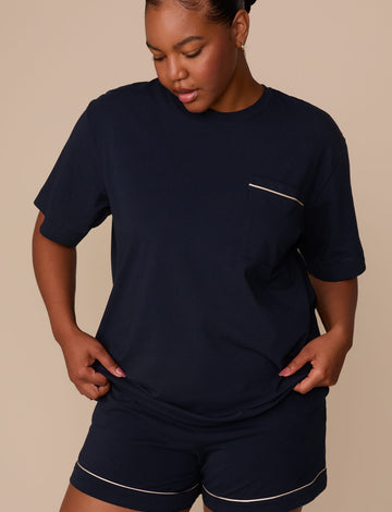 On a warm beige studio backdrop, Zarria wears the Perfect Pima Pajama T-Shirt in a deep navy color with matching pajama shorts. The angled front view highlights the relaxed fit, crew neckline, short sleeves, and chest pocket with contrasting piping detail. sizing: Laura wears size XL