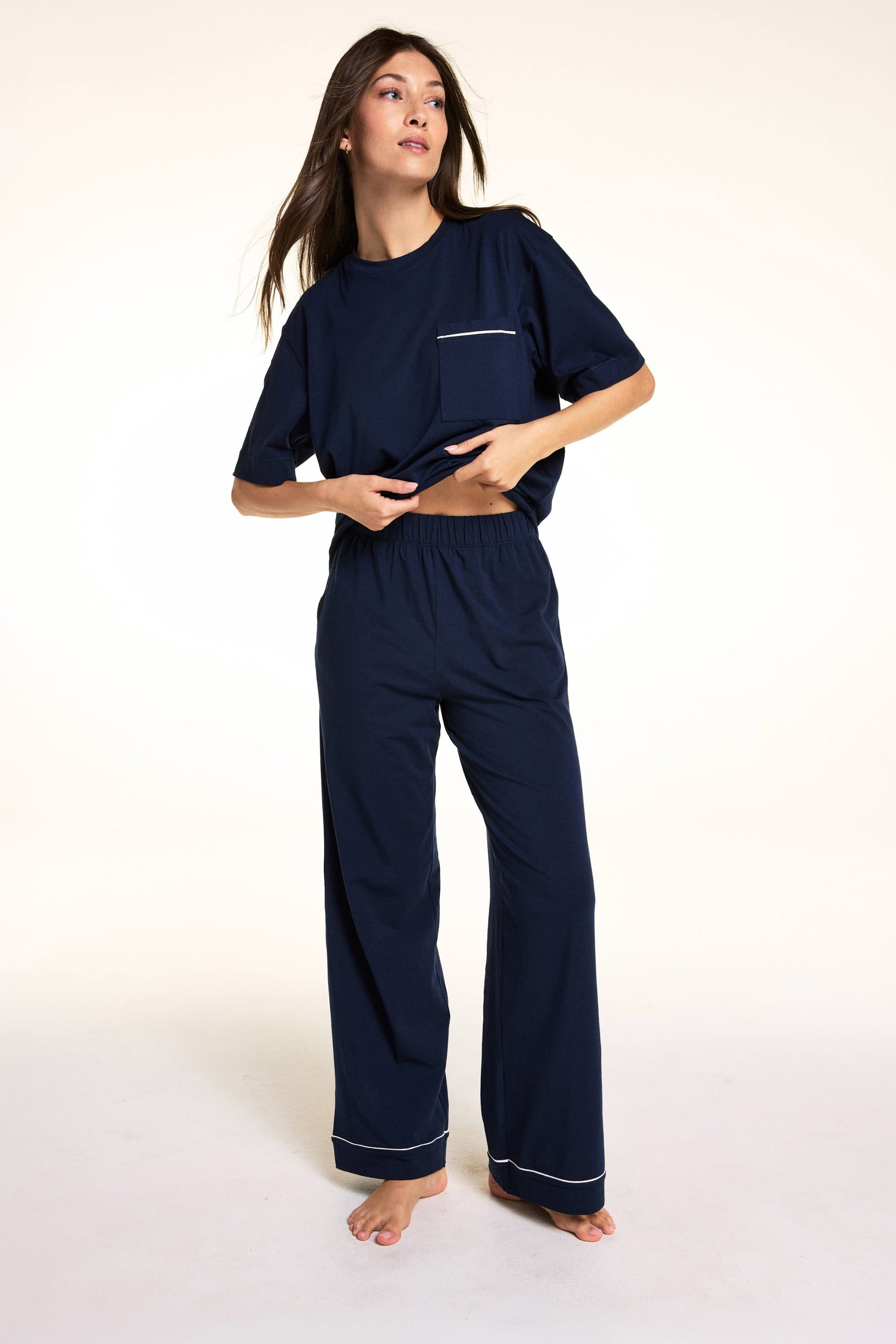 Against a neutral studio backdrop, Meagan wears the Perfect Pima Pant Set in Navy, lifting the hem of the relaxed short-sleeve sleep tee to show the elastic waistband and matching wide-leg lounge pants. sizing: Meagan wears size Small