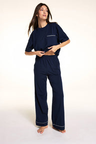 Against a neutral studio backdrop, Meagan wears the Perfect Pima Pant Set in Navy, lifting the hem of the relaxed short-sleeve sleep tee to show the elastic waistband and matching wide-leg lounge pants. sizing: Meagan wears size Small