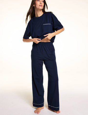 Against a neutral studio backdrop, Meagan wears the Perfect Pima Pant Set in Navy, lifting the hem of the relaxed short-sleeve sleep tee to show the elastic waistband and matching wide-leg lounge pants. sizing: Meagan wears size Small