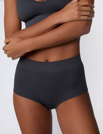 In a studio setting, Courtney wears the Ribbed Seamless Boyshort in Charcoal with a matching ribbed bralette. A close-up view highlights the high-rise waistband, boyshort silhouette, and ribbed texture.