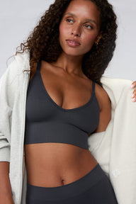 In a studio setting, Courtney wears the Ribbed Seamless Triangle Bralette in Charcoal with a matching ribbed bottom. A zip-up hoodie drapes off her shoulders, highlighting the ribbed texture, V-neckline, adjustable straps, and wide underband. sizing: Courtney wears size Small