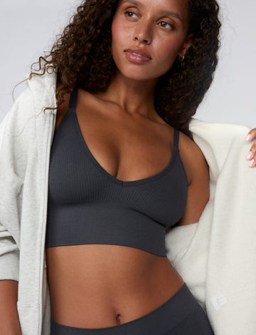 In a studio setting, Courtney wears the Ribbed Seamless Triangle Bralette in Charcoal with a matching ribbed bottom. A zip-up hoodie drapes off her shoulders, highlighting the ribbed texture, V-neckline, adjustable straps, and wide underband. sizing: Courtney wears size Small