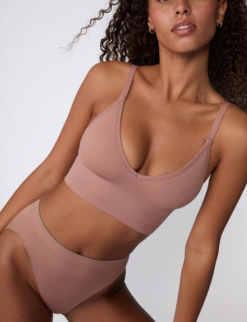 On a light gray studio backdrop, Courtney wears the Ribbed Seamless Triangle Bralette in Plum with matching underwear. The close-up angle highlights the deep V neckline, ribbed seamless fabric, and longline band. sizing: Courtney wears size Small