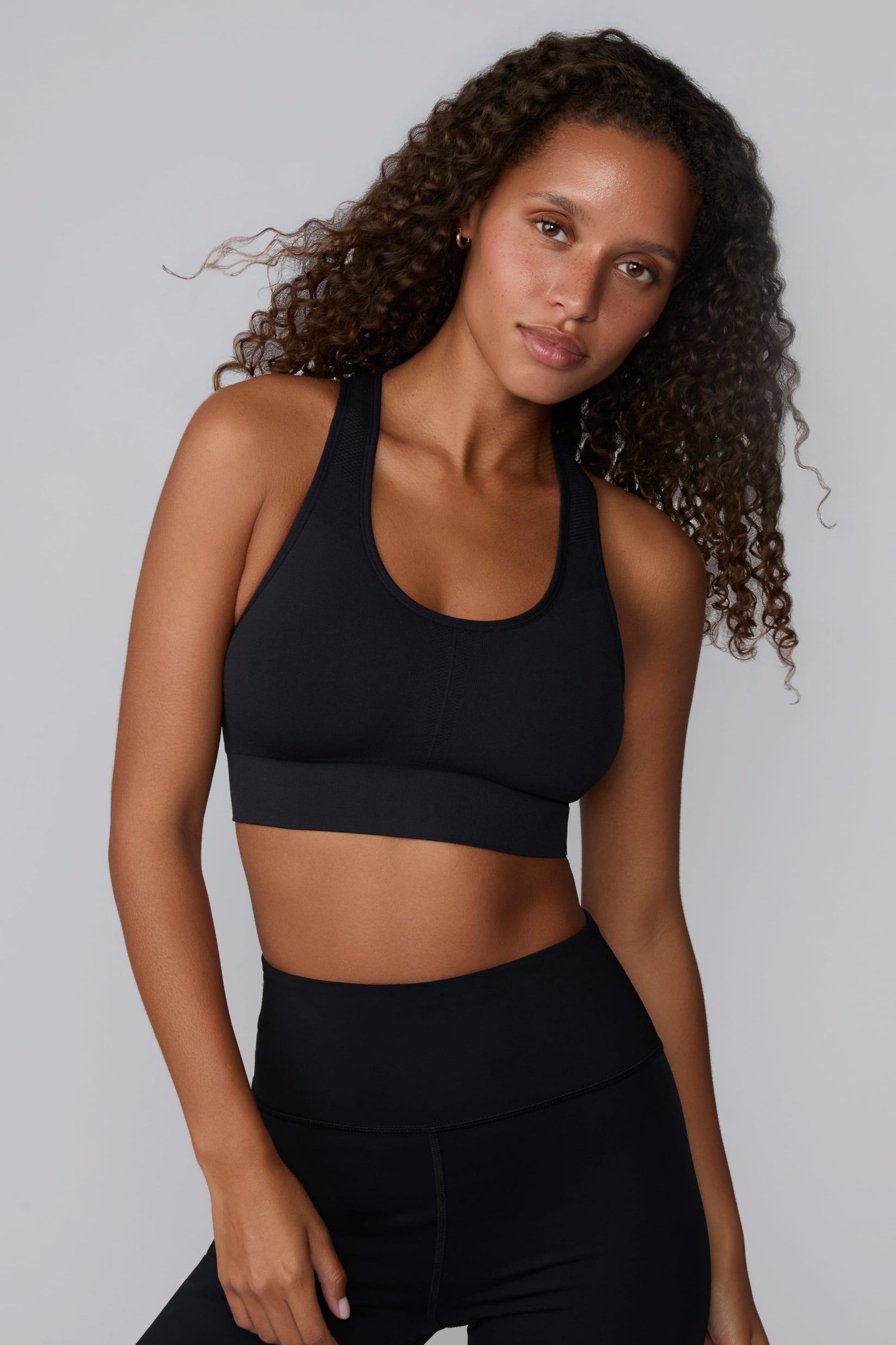 On a light gray studio backdrop, Courtney wears the Seamless Racerback Sports Bra in Black with matching bike shorts. The front-facing view highlights the scoop neckline, wide underband, racerback straps, and seamless knit construction.
 sizing: Courtney wears size Small