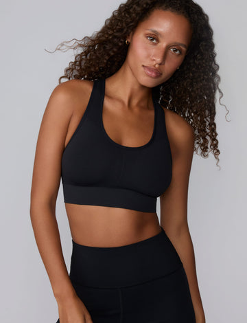 On a light gray studio backdrop, Courtney wears the Seamless Racerback Sports Bra in Black with matching bike shorts. The front-facing view highlights the scoop neckline, wide underband, racerback straps, and seamless knit construction.
 sizing: Courtney wears size Small