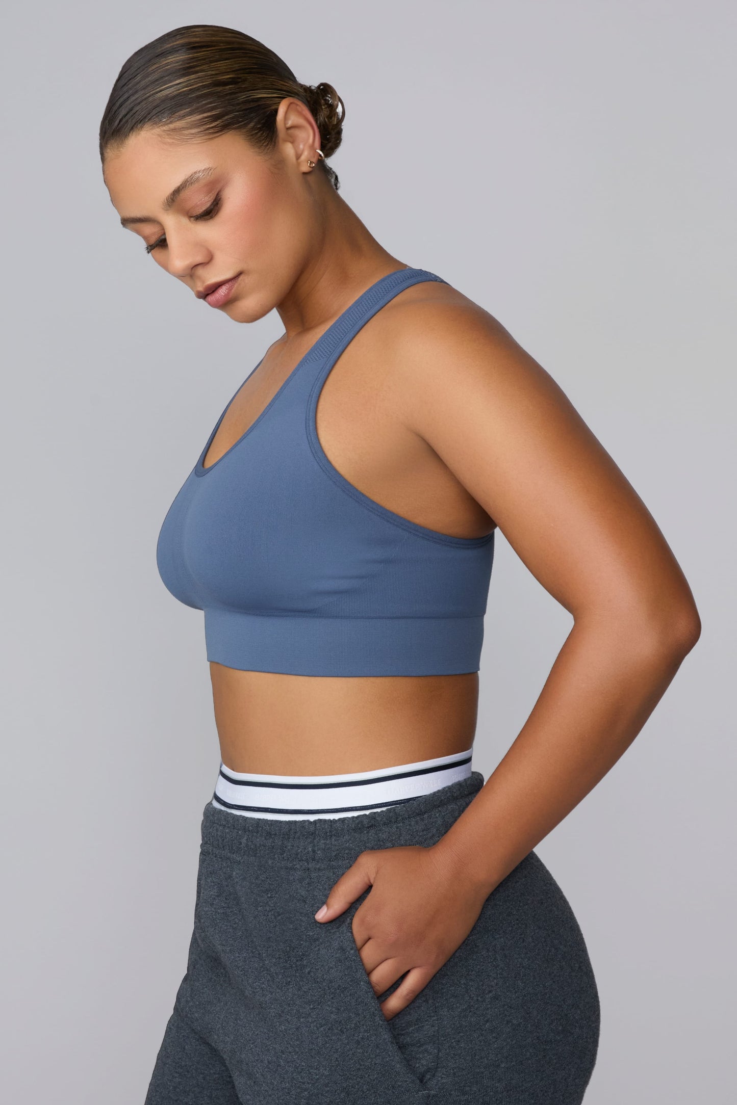 On a light gray studio backdrop, India wears the Seamless Racerback Sports Bra in Evernight with charcoal joggers. The side-profile view highlights the scoop neckline, wide underband, and smooth seamless knit construction. sizing: India wears size Large