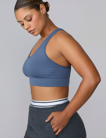 On a light gray studio backdrop, India wears the Seamless Racerback Sports Bra in Evernight with charcoal joggers. The side-profile view highlights the scoop neckline, wide underband, and smooth seamless knit construction. sizing: India wears size Large