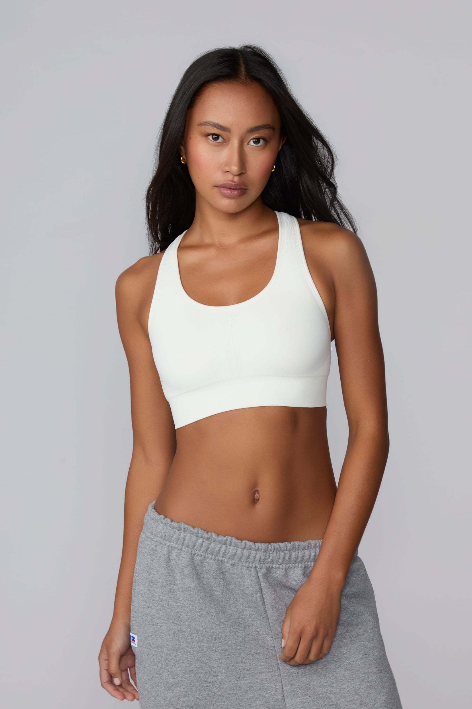 On a light gray studio backdrop, Amber wears the Seamless Racerback Sports Bra in White with gray sweatpants. The front-facing view highlights the scoop neckline, wide underband, racerback straps, and seamless knit construction. sizing: Laura wears size XS