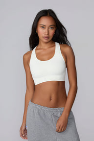 On a light gray studio backdrop, Amber wears the Seamless Racerback Sports Bra in White with gray sweatpants. The front-facing view highlights the scoop neckline, wide underband, racerback straps, and seamless knit construction. sizing: Laura wears size XS
