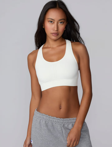 On a light gray studio backdrop, Amber wears the Seamless Racerback Sports Bra in White with gray sweatpants. The front-facing view highlights the scoop neckline, wide underband, racerback straps, and seamless knit construction. sizing: Laura wears size XS