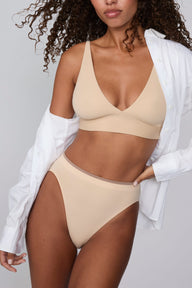 On a light gray studio backdrop, Courtney wears the Smooth Seamless Brief in Beige with the matching Smooth Seamless Bralette in Beige. An open white button-down shirt drapes off her shoulders, while the front view highlights the high-leg silhouette, smooth waistband with contrast trim, and comfortable coverage. sizing: Courtney wears size Small
