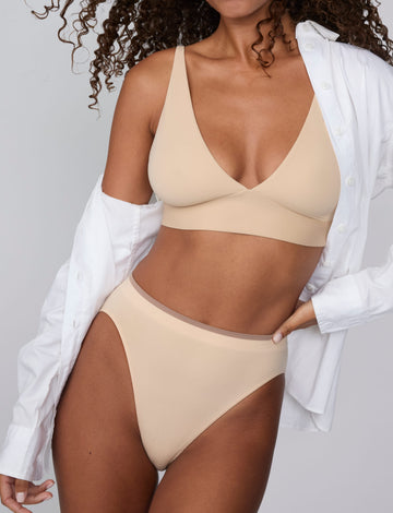 On a light gray studio backdrop, Courtney wears the Smooth Seamless Brief in Beige with the matching Smooth Seamless Bralette in Beige. An open white button-down shirt drapes off her shoulders, while the front view highlights the high-leg silhouette, smooth waistband with contrast trim, and comfortable coverage. sizing: Courtney wears size Small