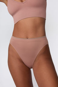 On a light gray studio backdrop, Courtney wears the Smooth Seamless Brief in Plum with the matching Seamless Scoop Bralette in Plum. A close-up front view highlights the high-leg silhouette, smooth fabric, and contrast trim waistband. sizing: Seamless wears size Small
