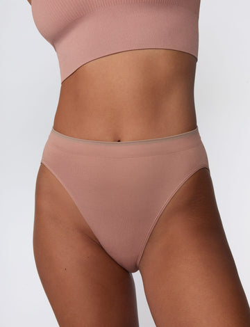 On a light gray studio backdrop, Courtney wears the Smooth Seamless Brief in Plum with the matching Seamless Scoop Bralette in Plum. A close-up front view highlights the high-leg silhouette, smooth fabric, and contrast trim waistband. sizing: Seamless wears size Small