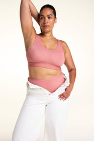 In a studio with a light neutral backdrop, Jennifer wears the Bliss Scoop Adjustable Bralette in Cinnamon with matching underwear and white pants worn open at the waist. She raises one arm to show the scoop neckline, wide straps, and smooth, wireless design. sizing: Jennifer wears size Large