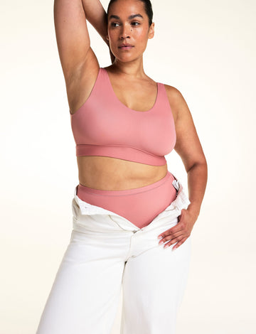 In a studio with a light neutral backdrop, Jennifer wears the Bliss Scoop Adjustable Bralette in Cinnamon with matching underwear and white pants worn open at the waist. She raises one arm to show the scoop neckline, wide straps, and smooth, wireless design. sizing: Jennifer wears size Large