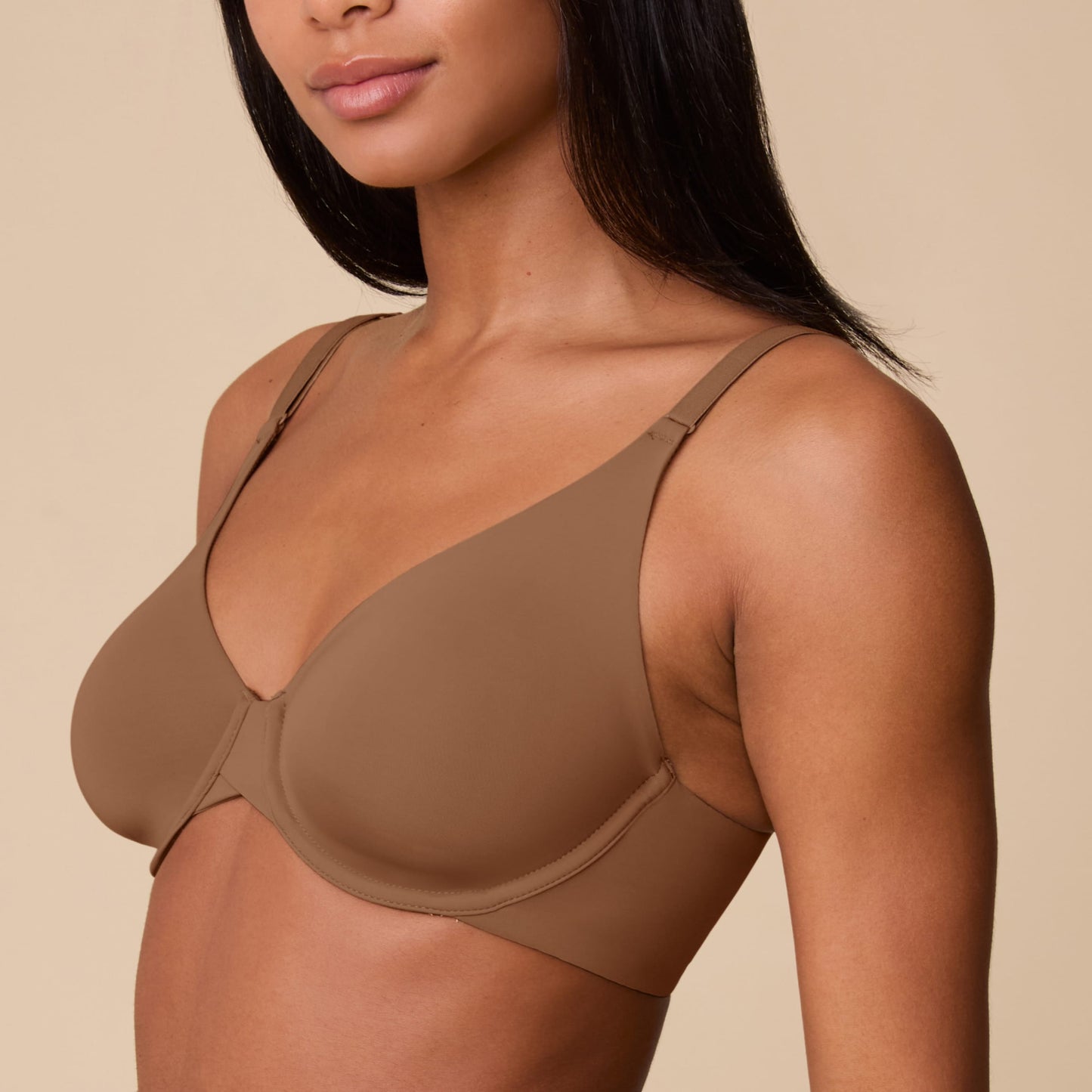 On a neutral beige backdrop, Ally wears the Base Unlined Bra in Brown, showing the unlined cups, underwire support, smooth microfiber fabric, and signature X-bridge at the center front.