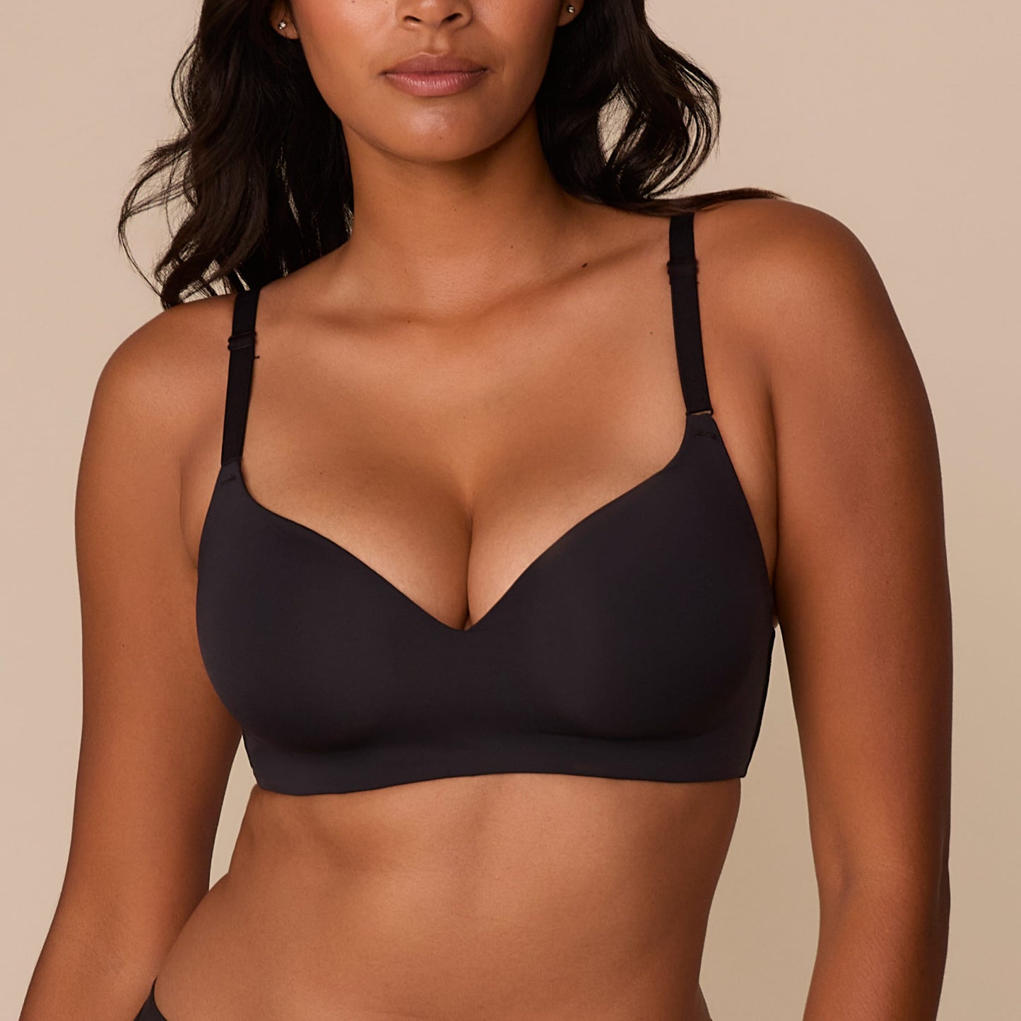On a neutral beige backdrop, a close-up of Victoria wearing the Base Wirefree T-Shirt Bra in Black showcases the smooth molded cups, wireless construction, and adjustable straps.