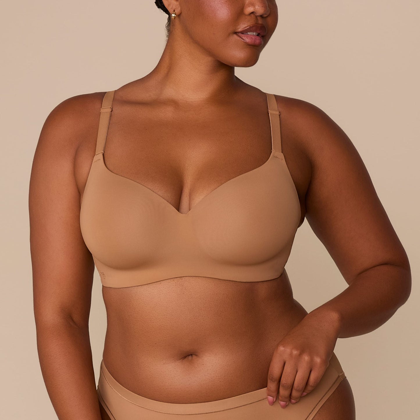 On a neutral beige backdrop, a close-up of Zarria wearing the Base Wirefree T-Shirt Bra in Tan showcases the smooth molded cups, wireless construction, and adjustable straps.