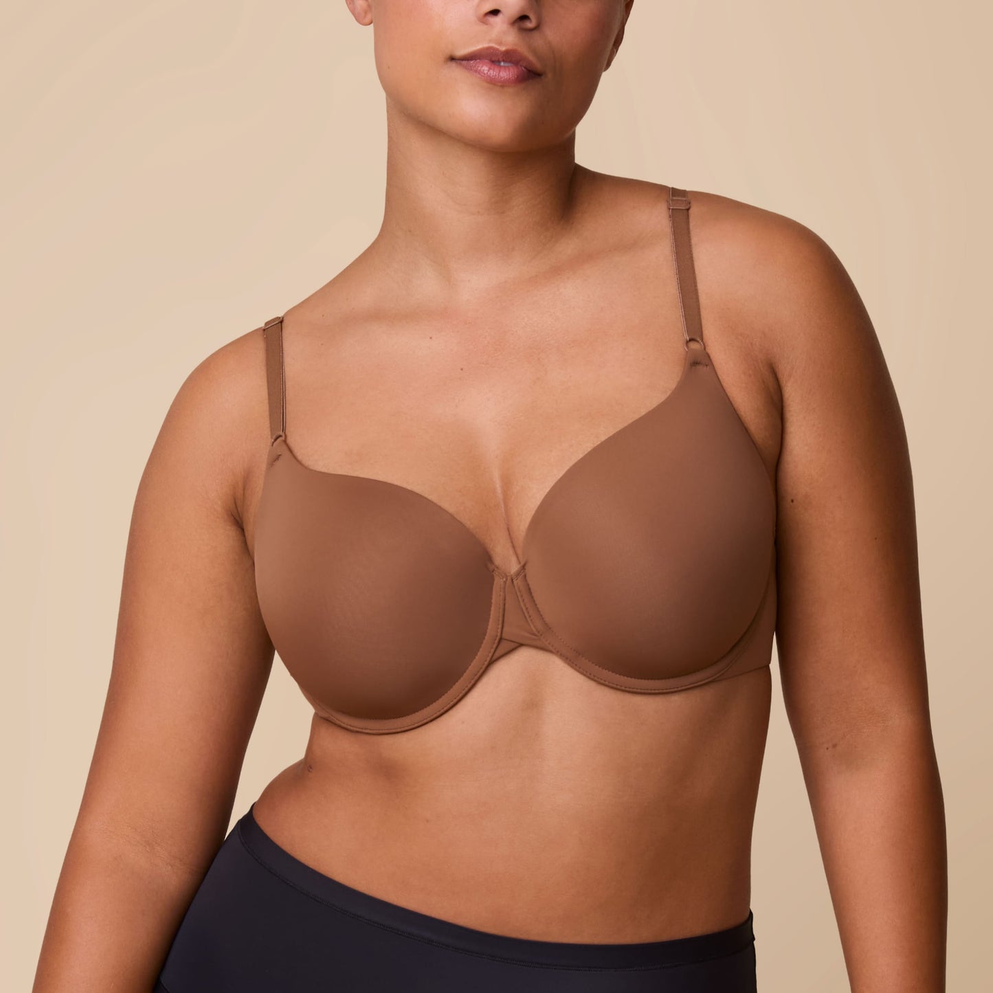 On a neutral beige backdrop, India wears the Base T-Shirt Bra in Brown, showing the smooth full-coverage cups, underwire support, and front-adjusting straps.