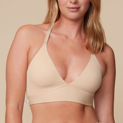 On a neutral beige backdrop, a close-up of Megan wearing the Bliss Lace Halterneck Bralette in Beige showcases the deep V-neckline, smooth cups, adjustable halterneck straps, and wide comfort band.