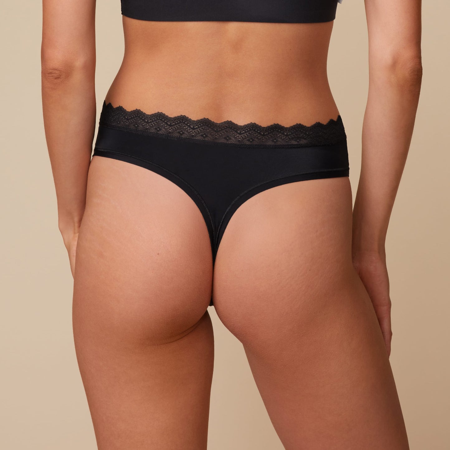 On a neutral beige backdrop, a back view of Anna wearing the Bliss Lace Highwaist Thong in Black showcases the thong back silhouette, smooth fabric, and lace-trim waistband.