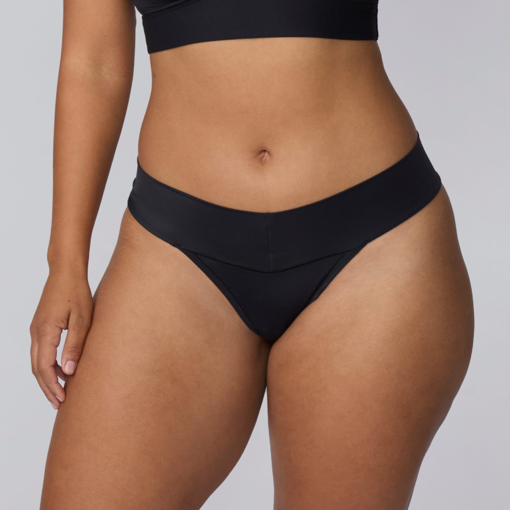 On a light gray studio backdrop, India wears the Bliss Low Rise Thong in Black. A close-up front view highlights the smooth waistband, low-rise fit, and minimal-coverage thong design.