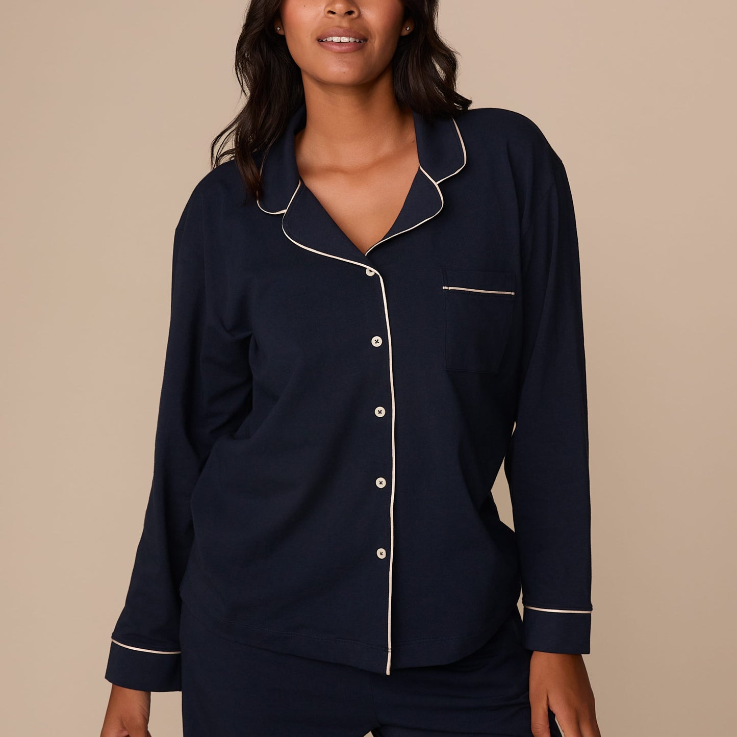 On a neutral studio backdrop, Victoria wears the Perfect Pima Pajama Long Sleeve Shirt in Navy with matching pajama pants. The cropped front view highlights the button-front closure, chest pocket, notched collar, and contrast piping details along the collar, placket, pocket, and cuffs.
