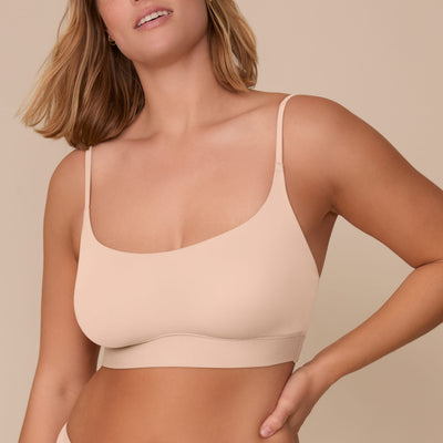 On a neutral beige backdrop, Sonnie wears the Bliss Scoop Adjustable Bralette in Beige, highlighting the scoop neckline, adjustable straps, and smooth underband while posing with one hand at her waist.