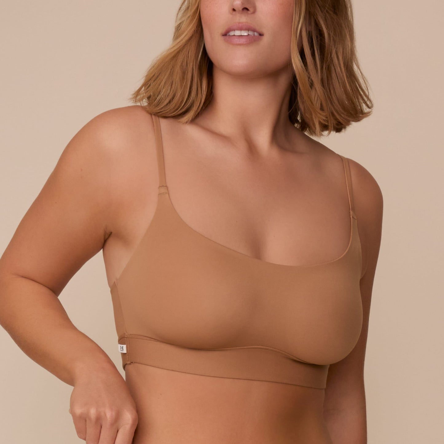 On a neutral beige backdrop, a close-up of Sonnie wearing the Bliss Scoop Adjustable Bralette in Tan highlights the scoop neckline, adjustable straps, and smooth fabric for a comfortable fit.