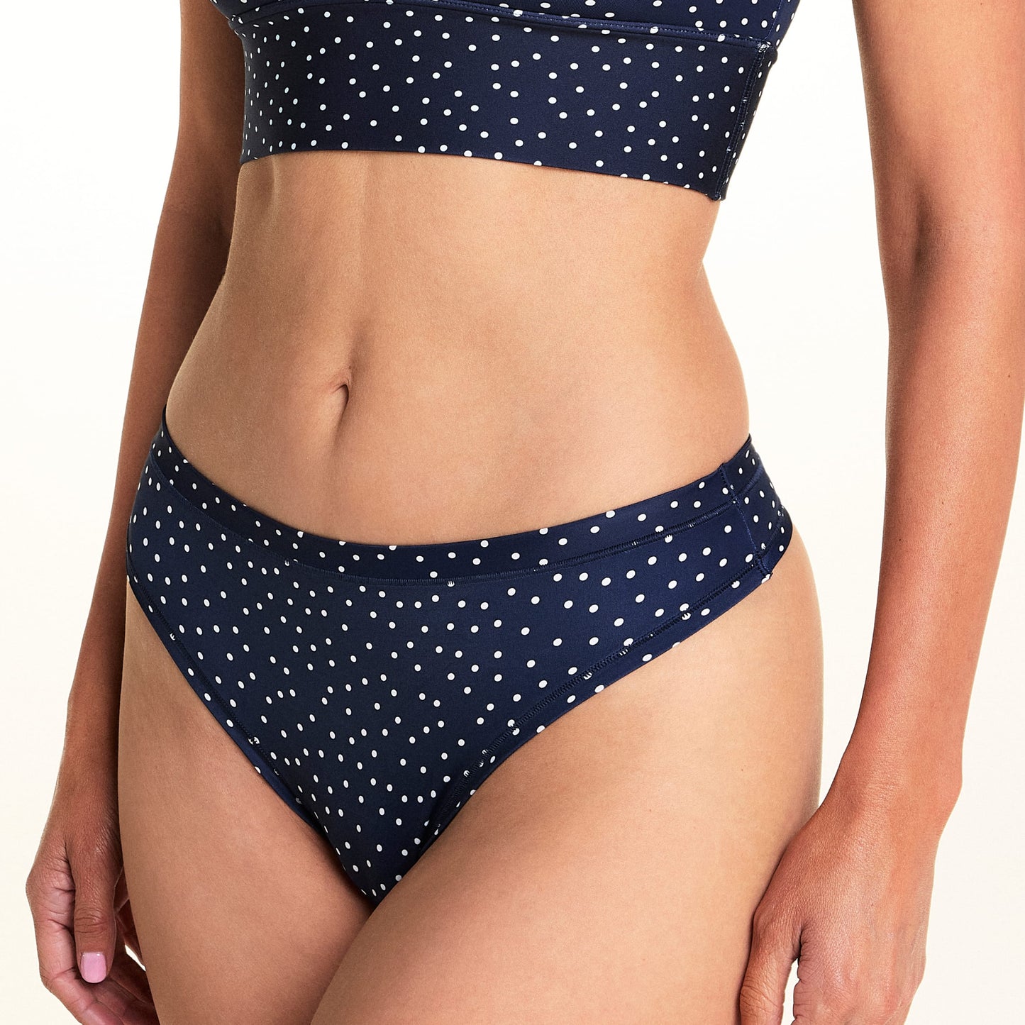 On a light studio backdrop, Meagan wears the Bliss Thong in Navy Dot. The cropped image highlights the low-rise waistband, thong silhouette, and allover white dot print.