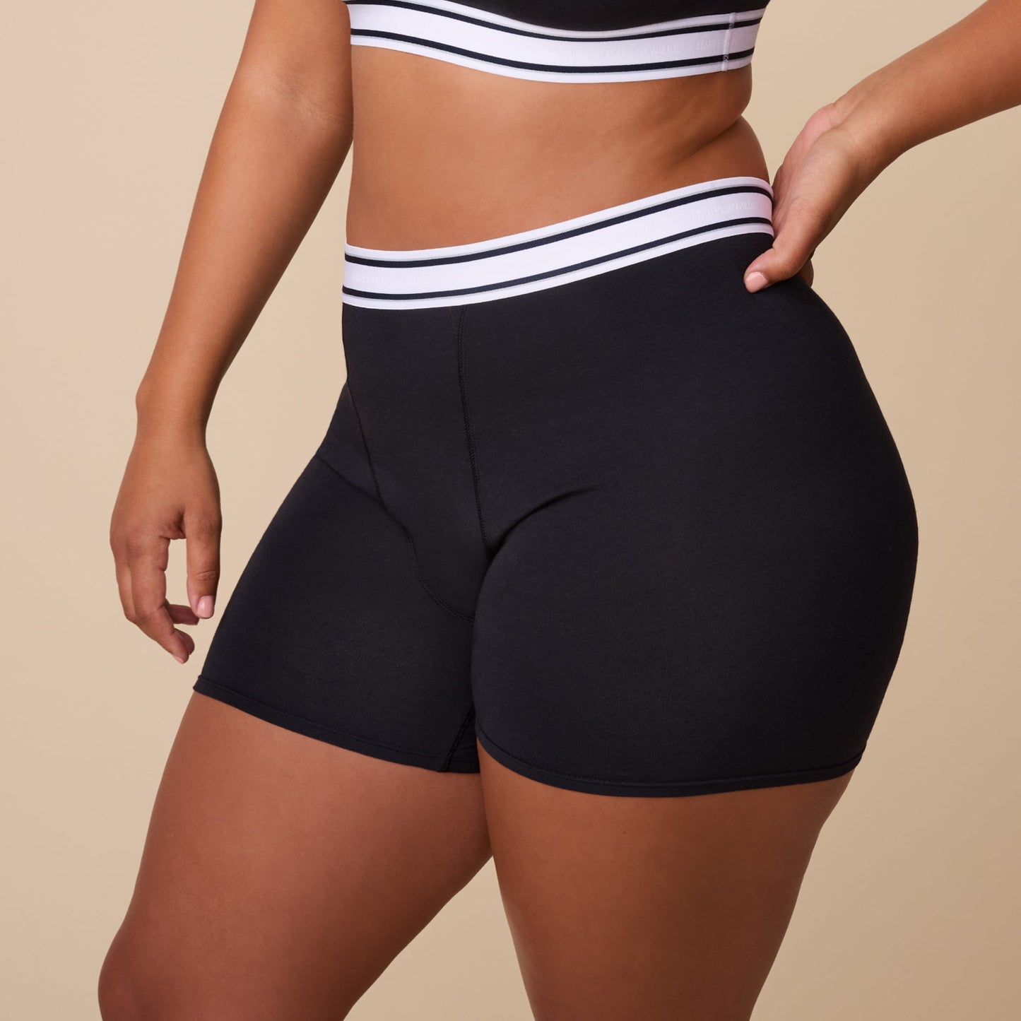 On a neutral studio backdrop, India wears the Cloud Cotton Retro Boxer Brief in Black. The close-up angled view highlights the full-coverage boxer brief silhouette, smooth cotton modal fabric, and retro-inspired logo waistband with contrasting stripe details.