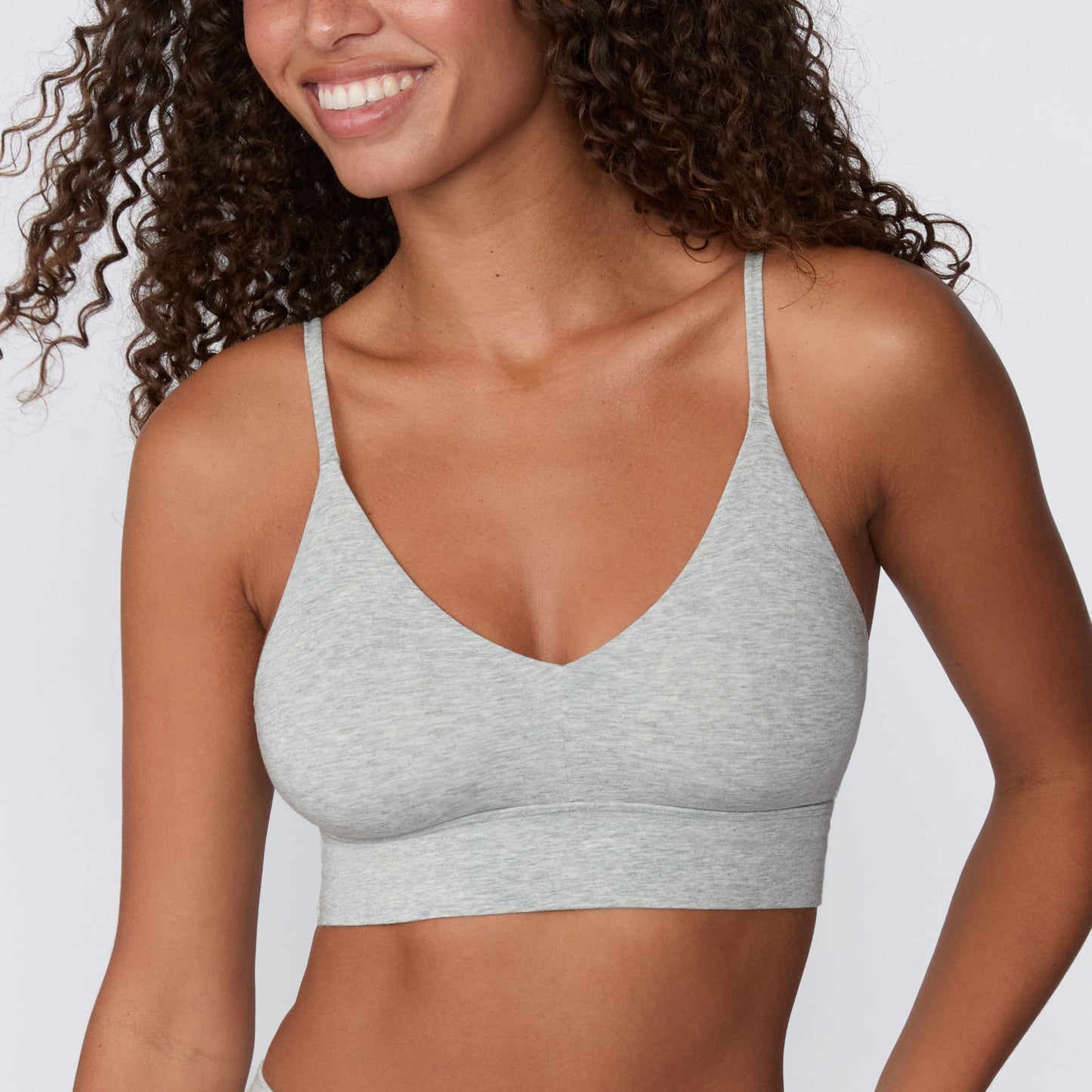 On a light gray studio backdrop, Courtney wears the Cloud Cotton Triangle Bralette in Heather Grey. The square-crop close-up highlights the soft cotton fabric, deep V-neckline, and thin adjustable straps as she smiles toward the camera.