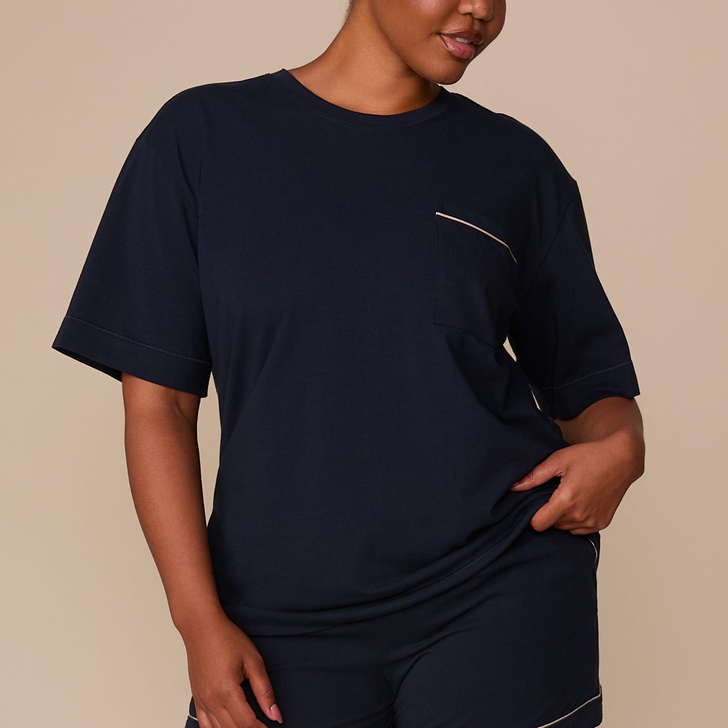 On a warm beige studio backdrop, Zarria wears the Perfect Pima Pajama T-Shirt in a deep navy color with matching pajama shorts. The cropped front view highlights the relaxed fit, crew neckline, short sleeves, and chest pocket with contrasting piping detail.