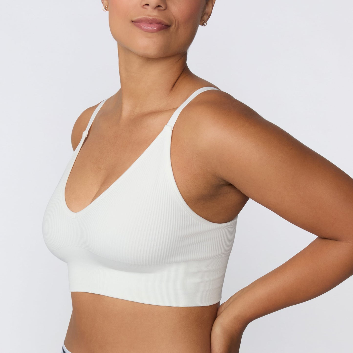 In a studio setting, India wears the Ribbed Seamless Triangle Bralette in White with gray lounge shorts. A cropped front-side view highlights the deep V-neckline, adjustable straps, ribbed texture, and wide supportive underband.