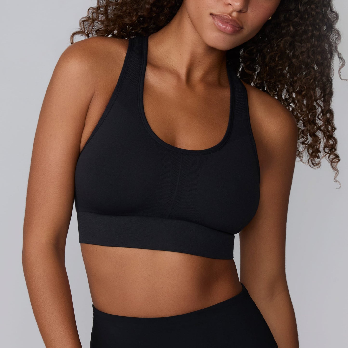 On a light gray studio backdrop, Courtney wears the Seamless Racerback Sports Bra in Black with matching bike shorts. The cropped front view highlights the scoop neckline, wide underband, racerback straps, and seamless knit construction.