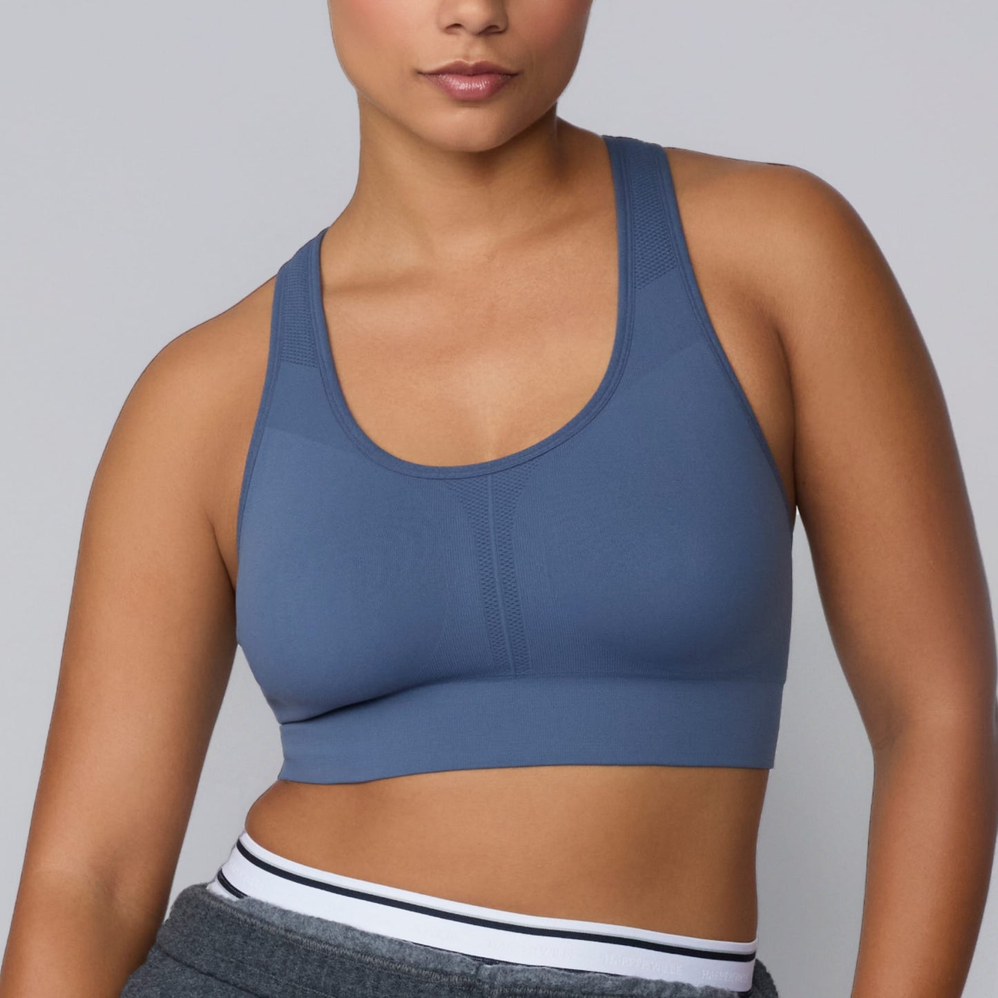 On a light gray studio backdrop, India wears the Seamless Racerback Sports Bra in Evernight. The cropped front view highlights the scoop neckline, wide straps, textured knit detailing, and supportive seamless construction.