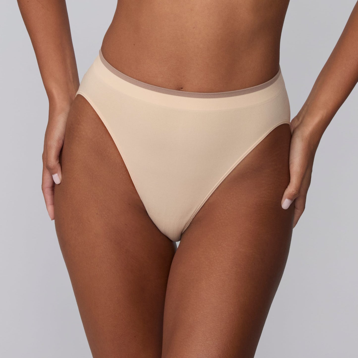 On a light gray studio backdrop, Courtney wears the Smooth Seamless Brief in Beige. A close-up front view highlights the high-leg cut, smooth finish, and contrast trim waistband.