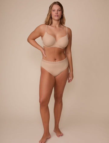 On a neutral studio backdrop, Sonnie wears the Base Lace T-Shirt Bra in Beige with matching underwear. The full-body view showcases the bra’s smooth cups, underwire construction, adjustable straps, and coordinating lace-trim underwear.