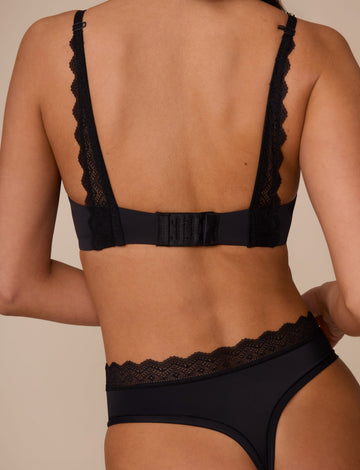 On a neutral studio backdrop, Marcela wears the Base Lace T-Shirt Bra in Black with matching underwear. The back view showcases the smooth band, adjustable straps, hook-and-eye closure, and lace detailing extending from the straps to the band.