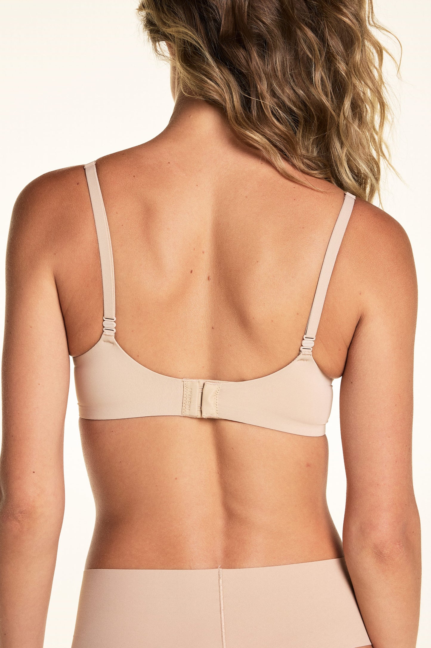 On a light studio backdrop, Alexis is shown from the back wearing the Base Picot T-Shirt Bra in Beige. The image highlights the adjustable straps and back hook-and-eye closure.
