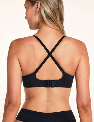 On a light studio backdrop, Meagan is shown from the back wearing the Base Picot T-Shirt Bra in Black with the straps in a racerback configuration. The image highlights the adjustable convertible straps and back hook-and-eye closure.