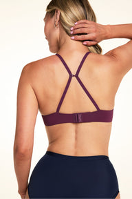 On a light studio backdrop, Meagan wears the Base T-Shirt Bra in Eggplant. The back view highlights the convertible J-hook straps, adjustable straps, and hook-and-eye closure.