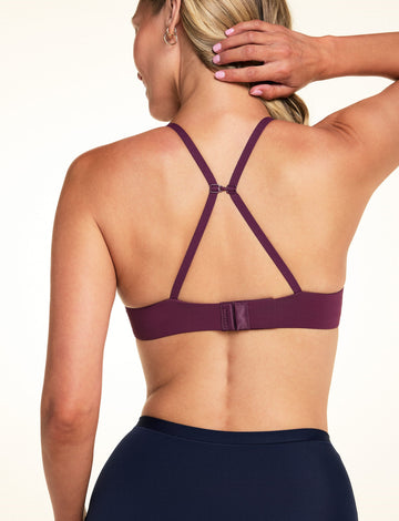 On a light studio backdrop, Meagan wears the Base T-Shirt Bra in Eggplant. The back view highlights the convertible J-hook straps, adjustable straps, and hook-and-eye closure.