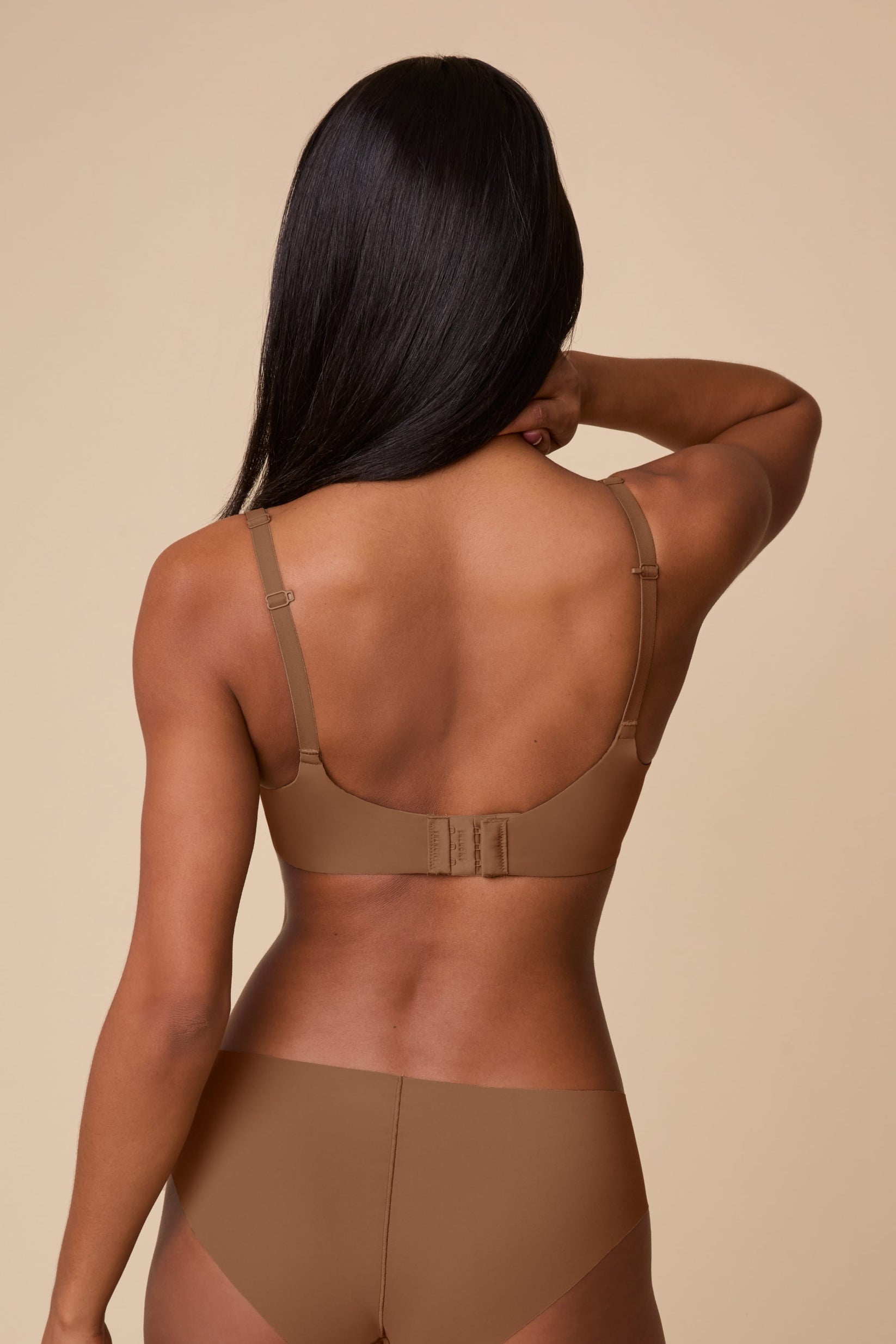 On a neutral beige backdrop, Ally wears the Base Unlined Bra in Brown, shown from the back to highlight the smooth band, adjustable straps, and hook-and-eye closure.