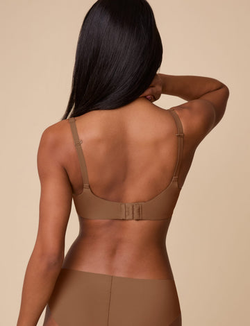 On a neutral beige backdrop, Ally wears the Base Unlined Bra in Brown, shown from the back to highlight the smooth band, adjustable straps, and hook-and-eye closure.